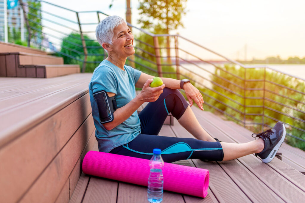 Smiling Senior Woman in Athletic Attire Sitting on Steps and Holding Apple_Senior Living Communities