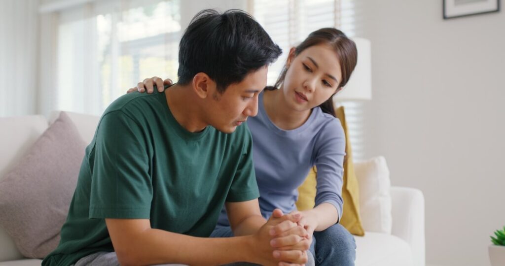 Adult Woman Puts Hand on Adult Man's Shoulder as they Sit on a Couch