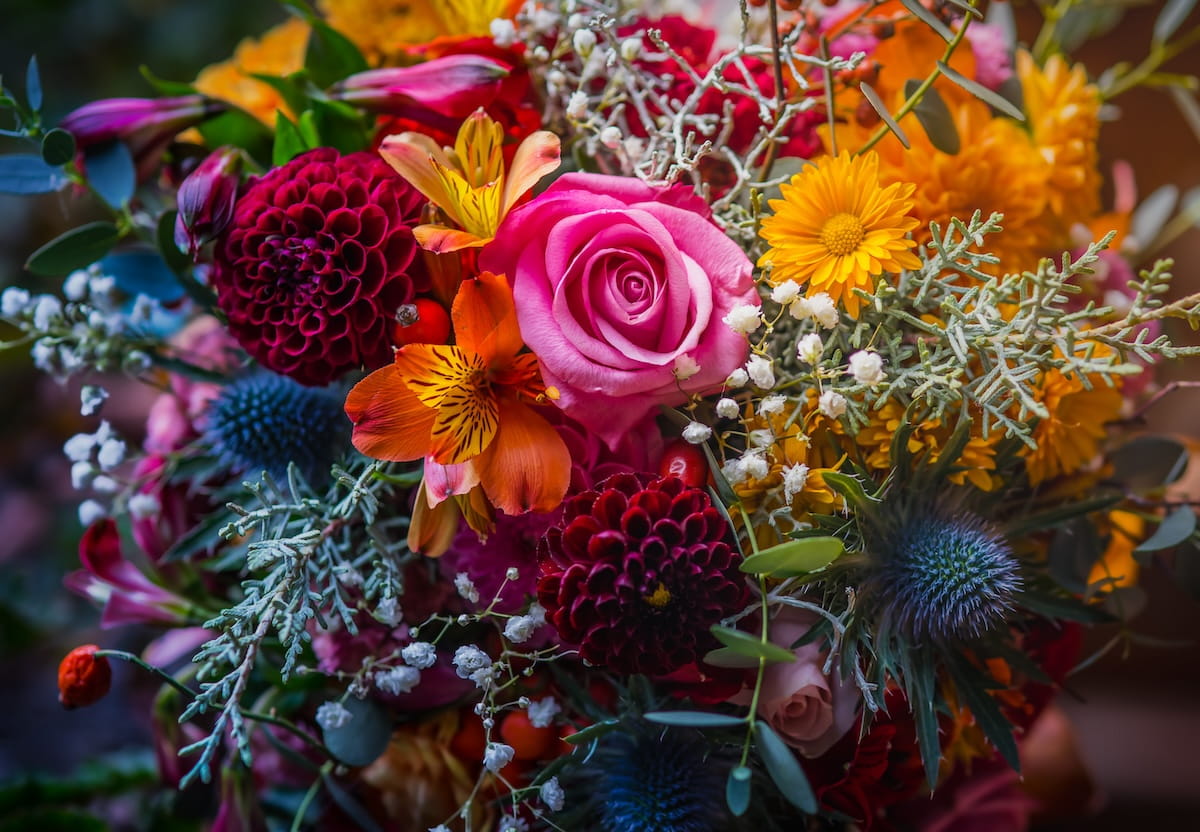 Vibrant bouquet with a pink rose, orange lilies, yellow daisies, blue thistles and white baby's breath in a lush arrangement.